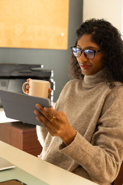 African American woman sitting at office desk wearing tan knit and glasses using tablet, copy space