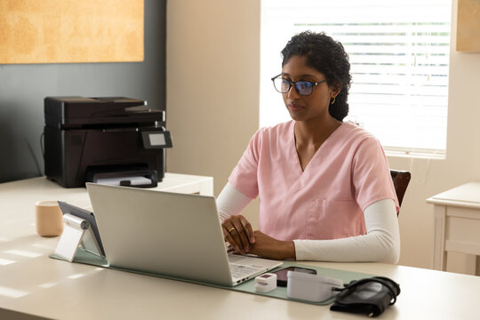Indian woman in pink scrubs sitting at clinic desk working on silver laptop with tablet, cuff