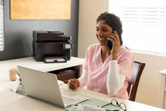 Adult African American female in pink scrubs sitting at clinic desk holding smartphone
