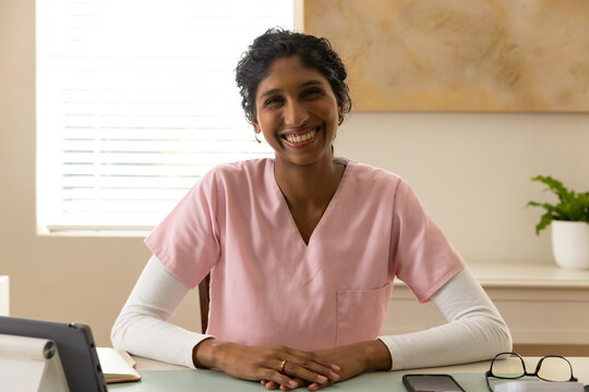 African American woman sitting at desk in consultation room wearing pink scrubs, using tablet
