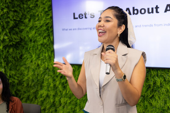 Adult woman presenting at meeting with living wall, holding mic, wearing blazer