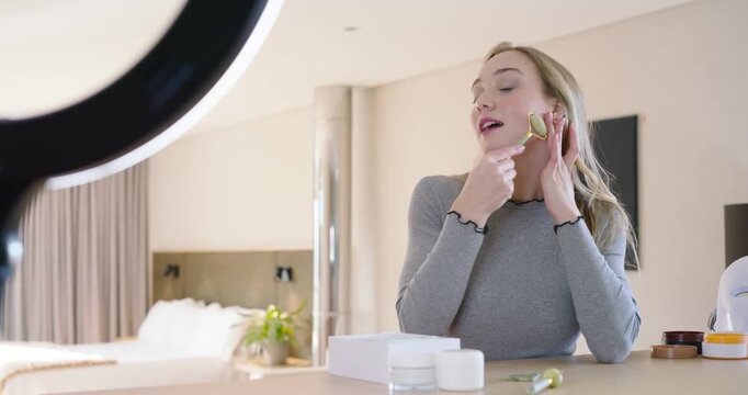 Adult female picking up green jade roller, showing rolling at vanity, ring light and skincare jars
