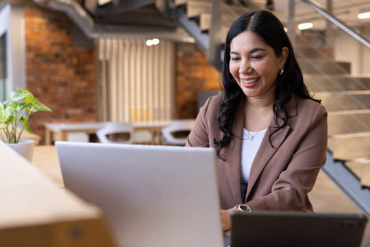 Woman smiling while working on silver laptop at open-plan office desk with brown blazer