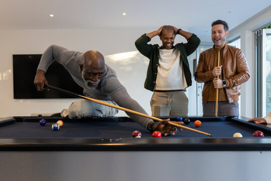 Diverse male friends lining up pool shot in home game room with cues, billiard balls
