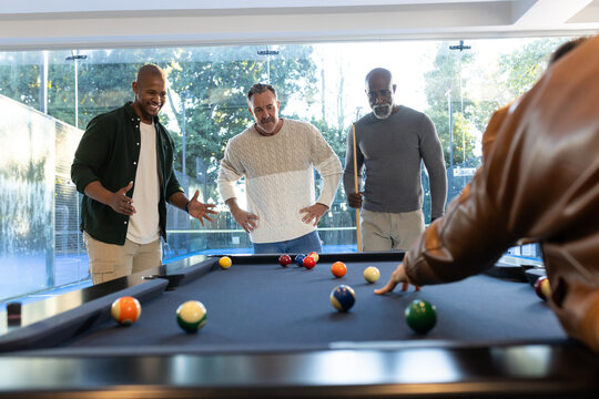 Diverse male friends wearing jackets playing pool at game room with dark felt table and cues