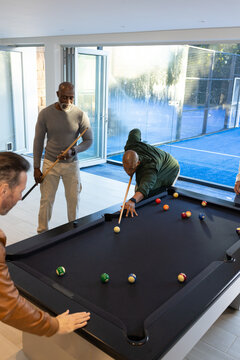 Diverse male friends playing billiards in modern recreation room around black-felt pool table