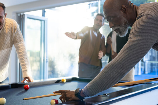 Diverse male friends playing pool in rec room, senior man lining up shot with cue stick