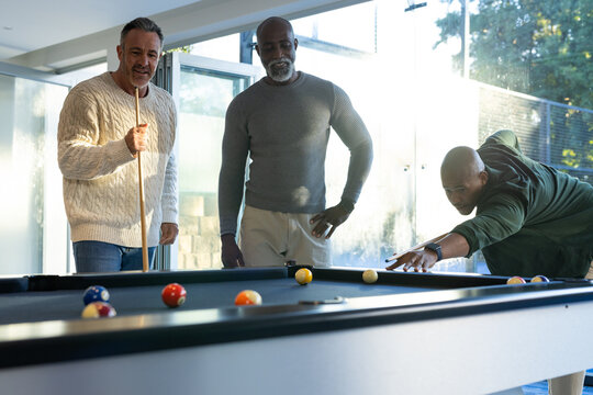 Diverse male friends leaning over pool table in modern recreation room, using cue sticks