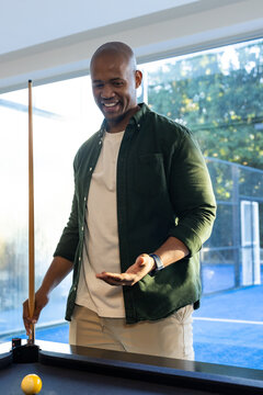 African-American man in green shirt standing by pool table holding cue near yellow ball by windows