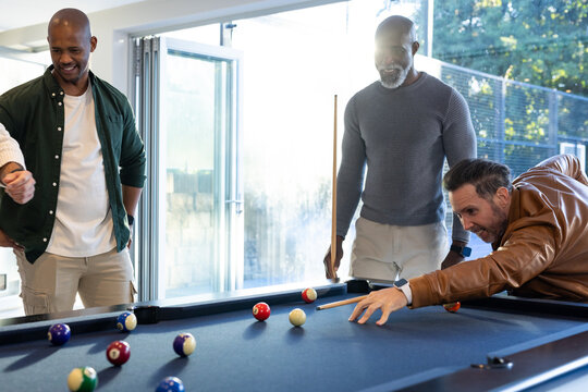 Diverse male friends leaning over navy-blue pool table at home, aiming cues at billiard balls