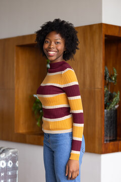 African American woman smiling while standing in lounge by wooden shelves wearing striped sweater