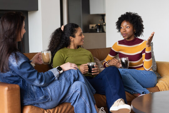 Diverse women chatting on brown leather sofa at home wearing denim and knit sweaters holding mugs