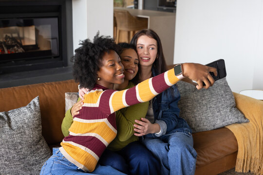 Diverse female friends sitting on brown leather sofa hugging and taking selfie with smartphone