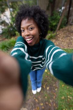 African woman taking selfie on garden path, smiling, holding smartphone, wearing green sweater