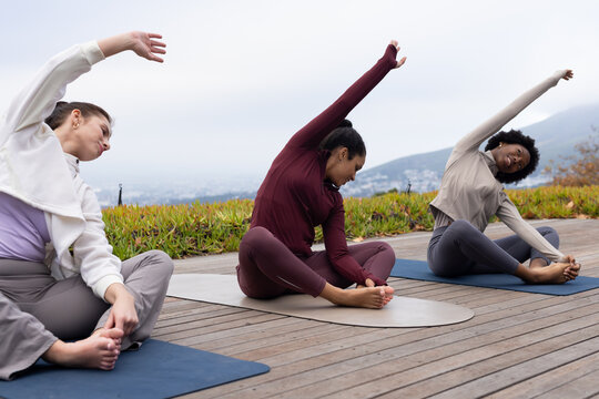 Diverse female friends in sportswear practicing seated side-stretch yoga on deck with yoga mats