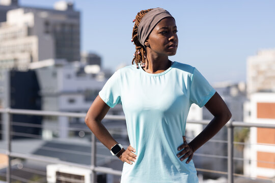 African woman standing on rooftop wearing blue athletic tee, headscarf, smartwatch by railing