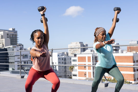Two adult African women performing weighted squats on rooftop terrace holding hex dumbbells