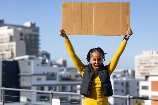 Woman holding blank cardboard sign and shouting on rooftop terrace wearing yellow top, braided hair