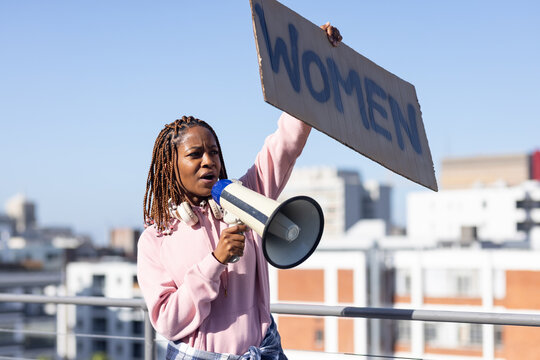 Adult African American woman holding WOMEN sign speaking into megaphone on roof in pink hoodie