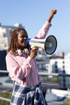 African woman standing on rooftop holding white megaphone and raising fist, wearing pink hoodie