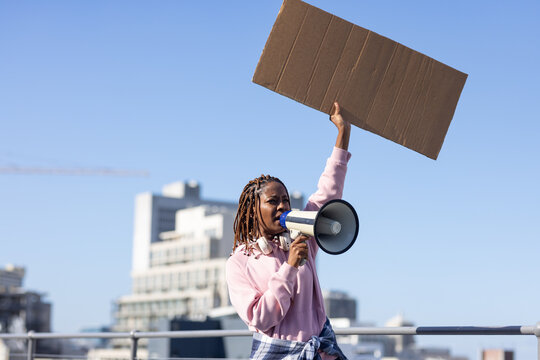 Woman wearing pink hoodie with headset holding megaphone and raising cardboard sign on roof railing