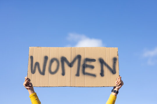 African woman holding corrugated placard reading WOMEN against bright blue sky, yellow sleeve