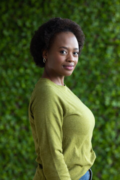 Adult African woman posing in 3 or 4 profile by hedge in light-green sweater, jeans, hoops, belt