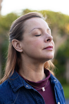 Female standing with eyes closed tilting head upward in garden wearing denim jacket and necklaces