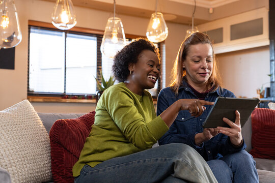 Diverse female friends sitting on couch by window, pointing at tablet with dark case under pendants