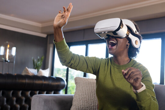 Adult woman sitting on sofa at home wearing green sweater and white VR headset, reaching forward