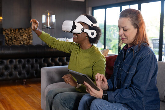 Female friends sitting on gray sofa at home left wearing green sweater using VR headset, tablet