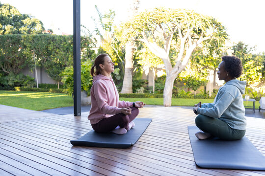 Diverse women friends in hoodies, leggings sitting on wooden deck meditating on yoga mats