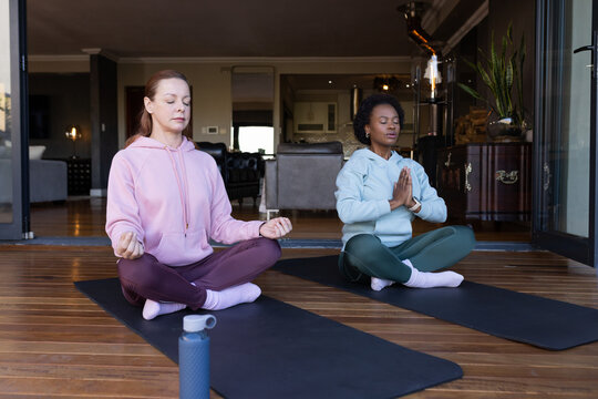 Diverse female friends meditating on two black mats on wooden deck wearing hoodies with teal bottle
