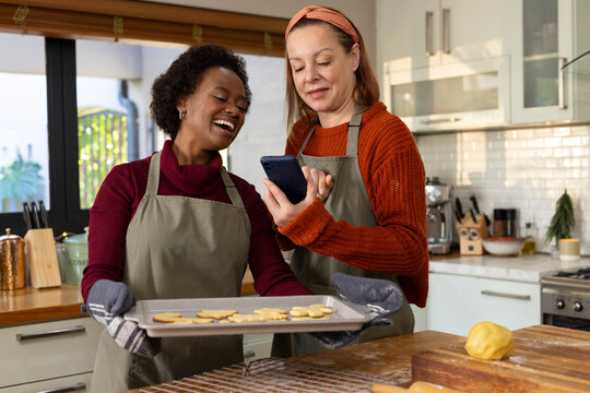 Diverse female friends laughing, holding cookie tray, wearing aprons and showing phone at counter