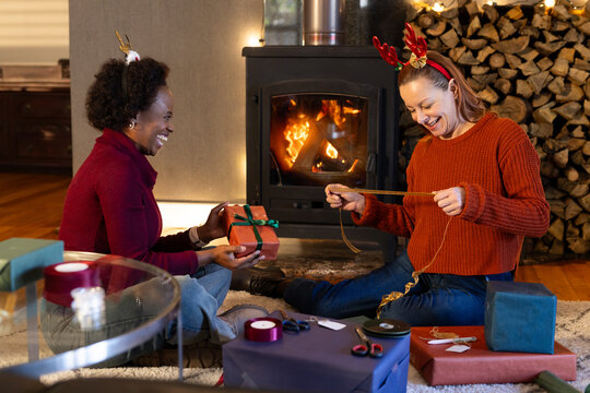 Diverse female friends sitting on rug at home wrapping gifts by wood-burning stove with ribbons