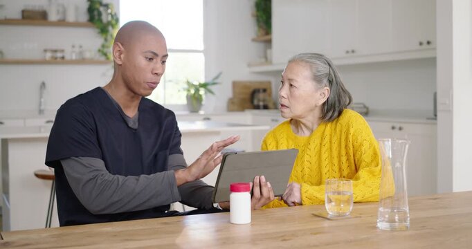 African American aide handing tablet pouring pills and showing dose to Asian senior woman at table