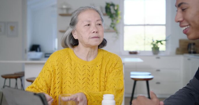 Diverse aide guiding Senior Asian after drinking at kitchen table on tablet checking pill bottle