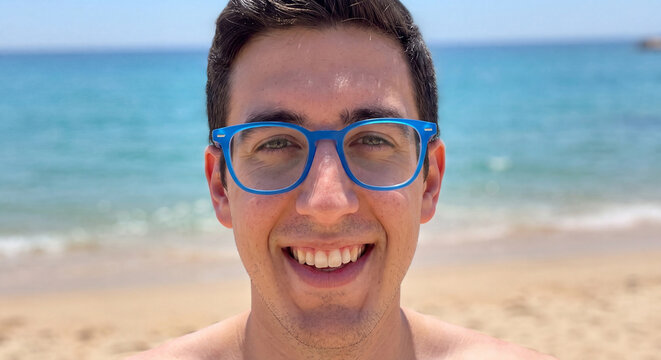 Cheerful young man wearing bright blue glasses smiling broadly while standing on a sunny seaside beach with ocean waters in background