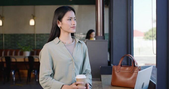 Mid-adult Asian woman sipping coffee cup while pausing laptop work at cafe window and smiling