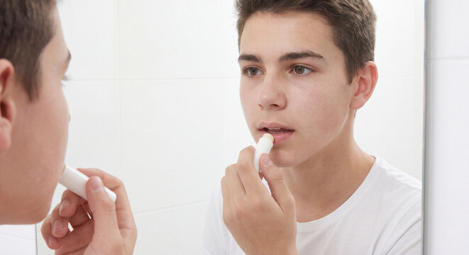 Young teenage boy applies a protective lip balm stick to his lips while looking into the bathroom mirror during a healthy male grooming and chapped lips hydration routine.