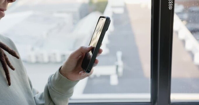 Non-binary adult green top adjusting, swiping dark-case phone, cutting glare at window, copy space