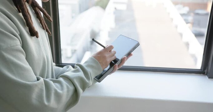 African American nonbinary in hoodie using stylus on tablet drawing at window, tilting, copy space