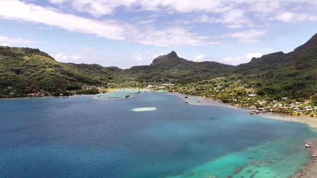 Aerial drone shot descending over the deep blue waters of Vaitape Bay, showcasing the central harbor town and lush tropical volcanic ridges of Bora Bora