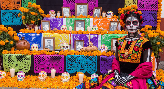 A woman painted in Day of the Dead Catrina makeup wearing traditional dress while sitting before a colorful ofrenda altar with marigolds