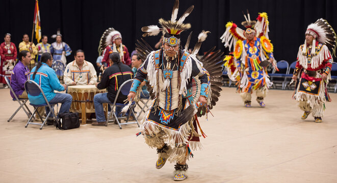 Native American dancers in ornate ceremonial feathered regalia performing traditional ritual dance steps on dirt ground during outdoor powwow