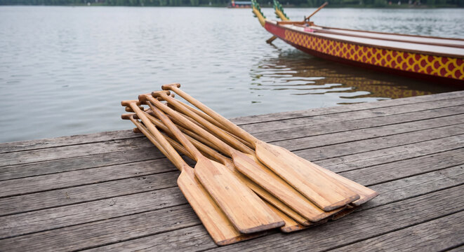 Neatly arranged wooden paddles lie on dock planks in foreground while traditional dragon boat rests along waterfront before water race
