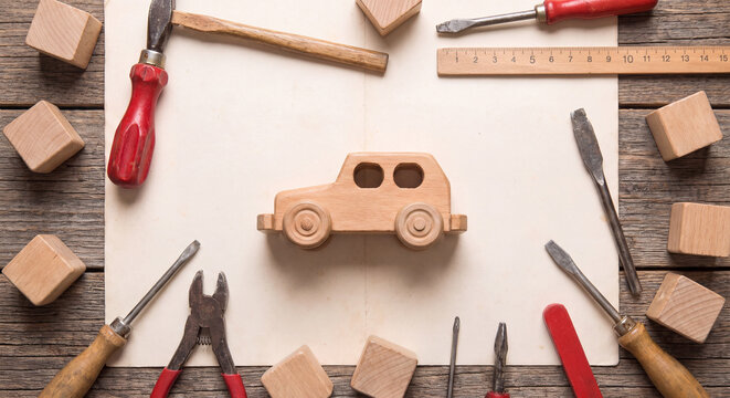 A craftsman wooden workbench flat lay displaying handmade wooden toy car surrounded by vintage chisels saw hammer and carving tools.
