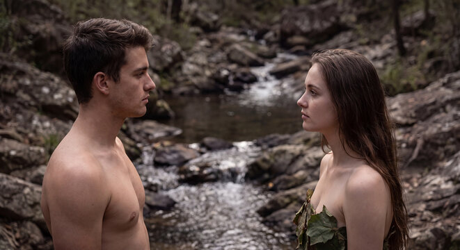 Portrayal of a young primitive prehistoric man and woman dressed in raw natural garments standing close together beside a rocky outdoor riverbank