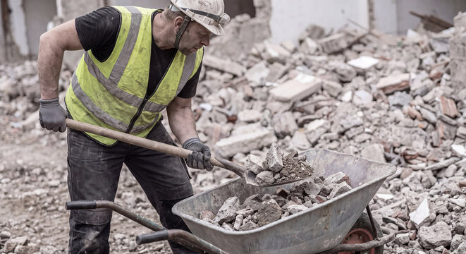 Construction worker in reflective safety vest using a metal shovel to load broken concrete pieces and gravel into a wheelbarrow