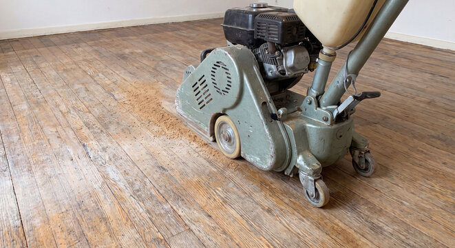 A heavy-duty electric belt sander machine sits stationed on a damaged domestic wooden parquet floor during an interior home renovation and refinishing project.